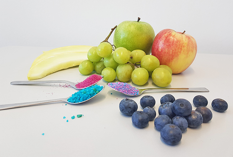 Drei Teelöffel mit buntem Badesalz, die zwischen Obst liegen.